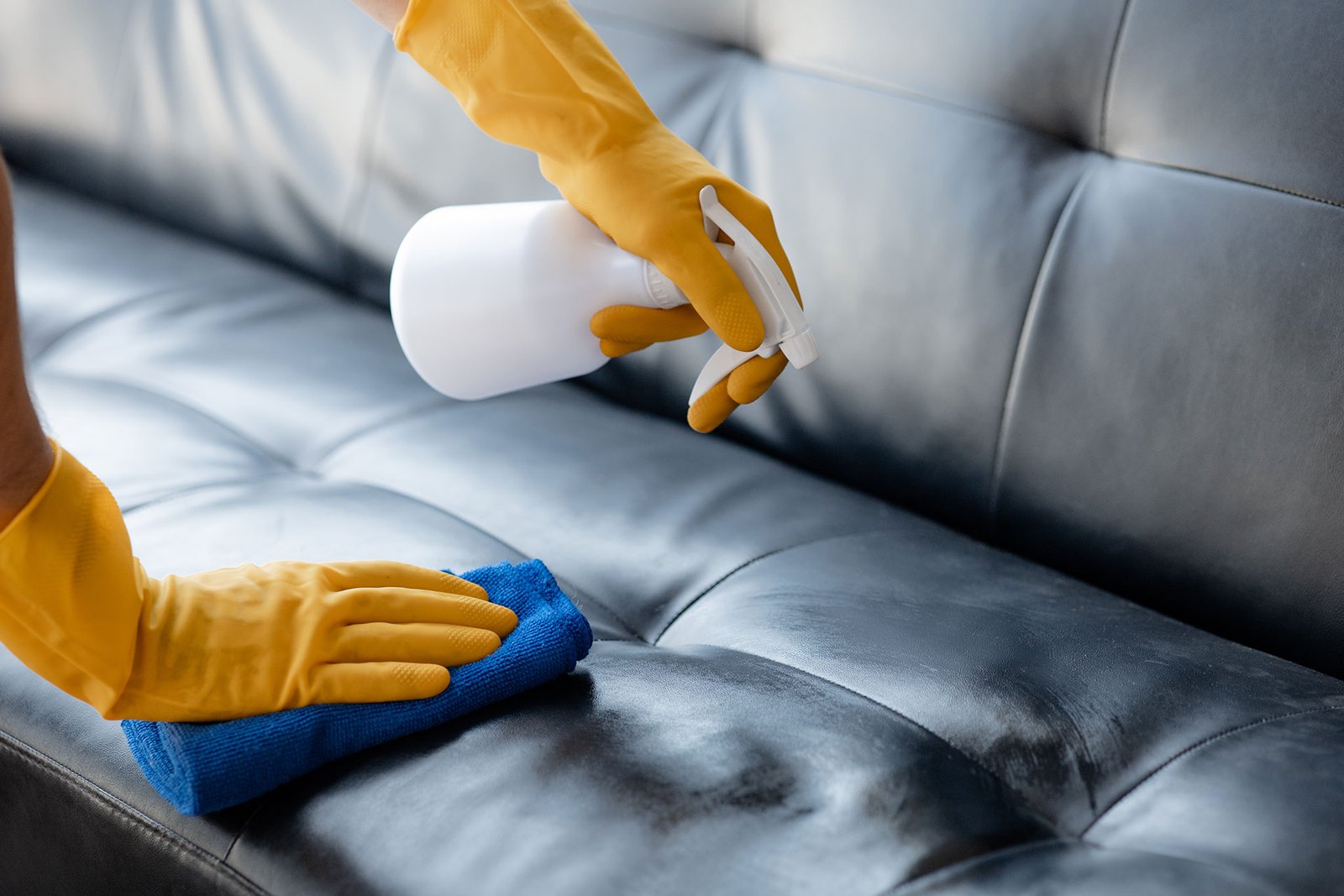 Hands in gloves cleaning black leather sofa with spray bottle and blue cloth.