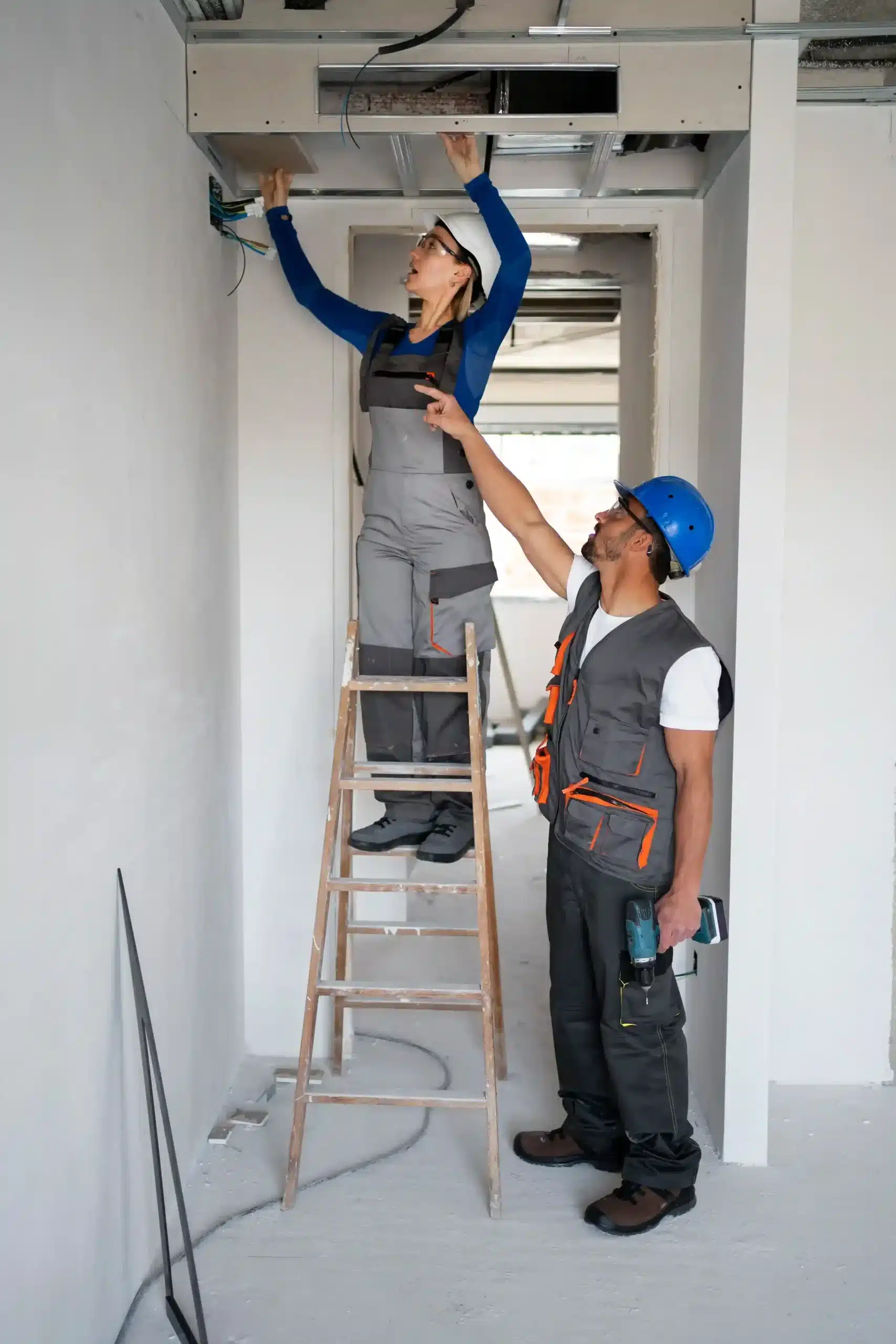 Two construction workers, one on a ladder and one on the ground, work together in a construction space, wearing helmets and safety gear.