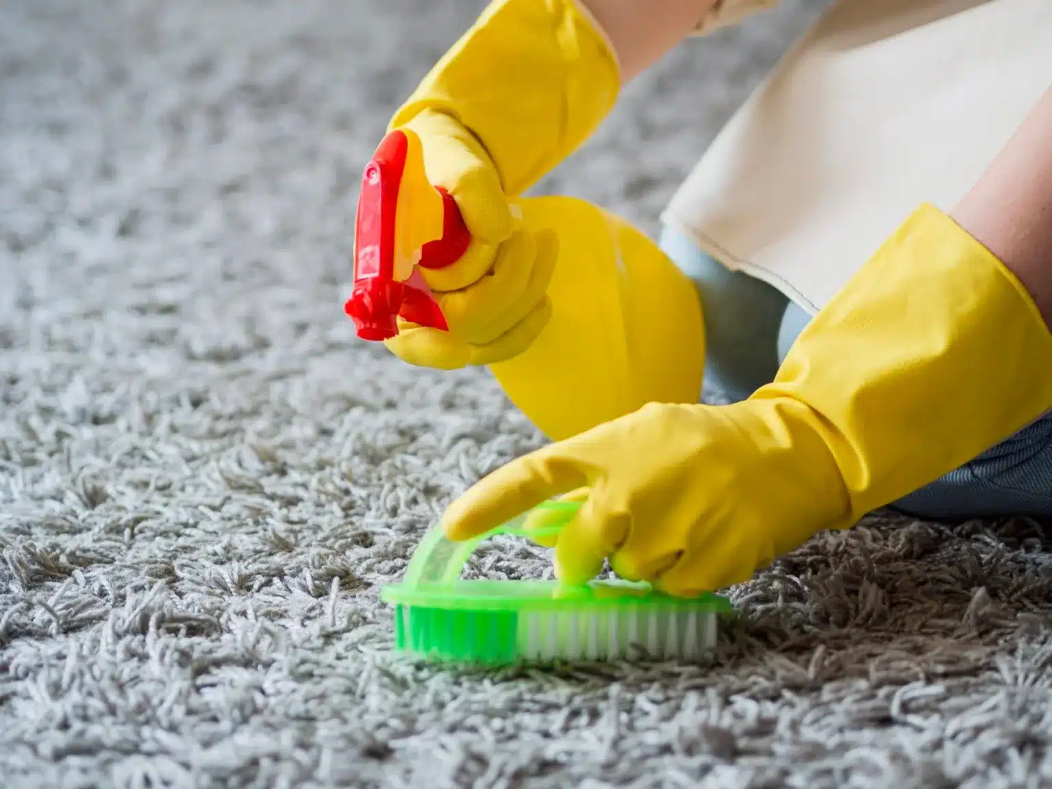 A person wearing yellow cleaning gloves sprays cleaner and scrubs a gray carpet with a green brush.