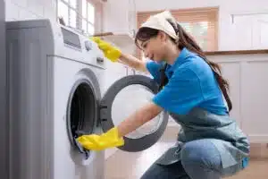 Woman wearing gloves and apron cleaning the inside of a front-load washing machine in a modern kitchen.