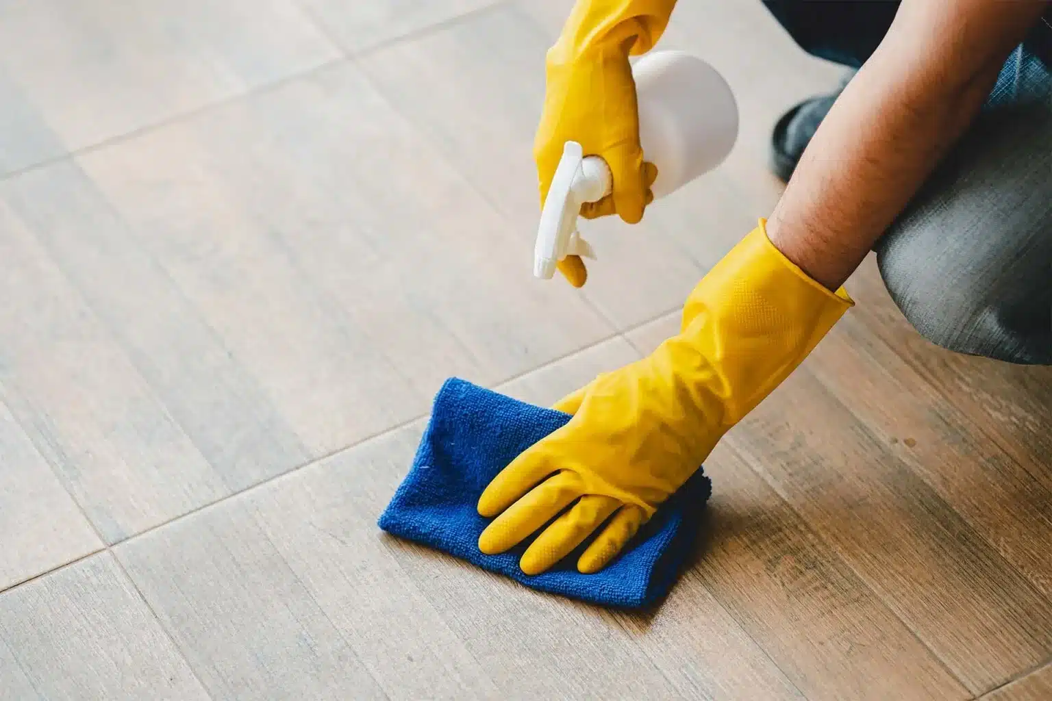 Person wearing yellow gloves cleaning tiled floor with spray bottle and blue cloth.