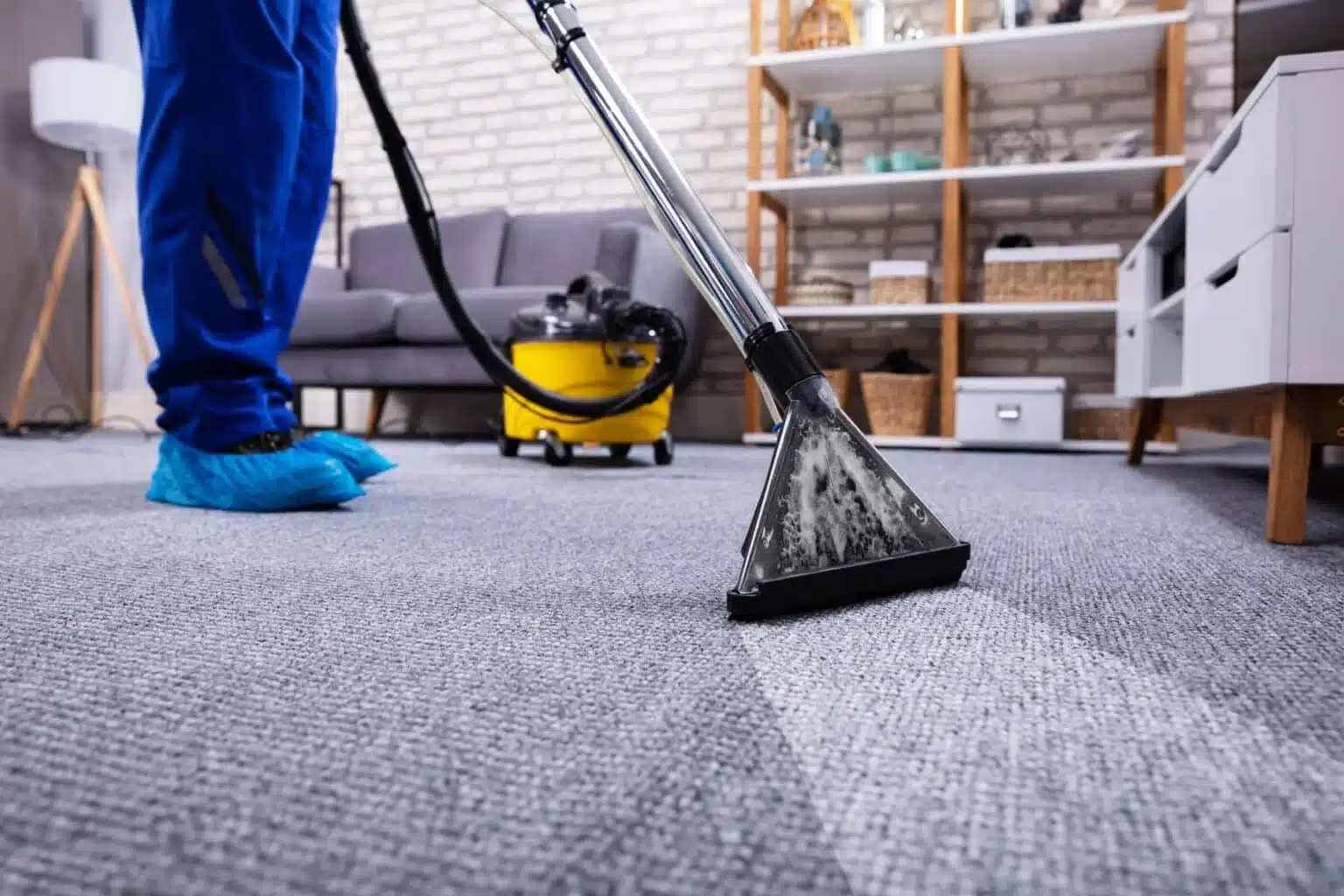 A cleaner using an industrial carpet cleaning machine on a grey carpet in a modern living room with a sofa and shelves in the background.