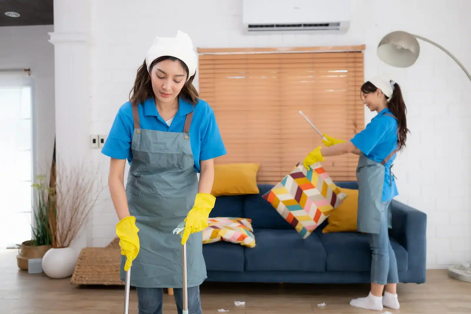 Two women wearing aprons and gloves cleaning a modern living room—one mopping the floor and the other arranging pillows on a blue sofa.