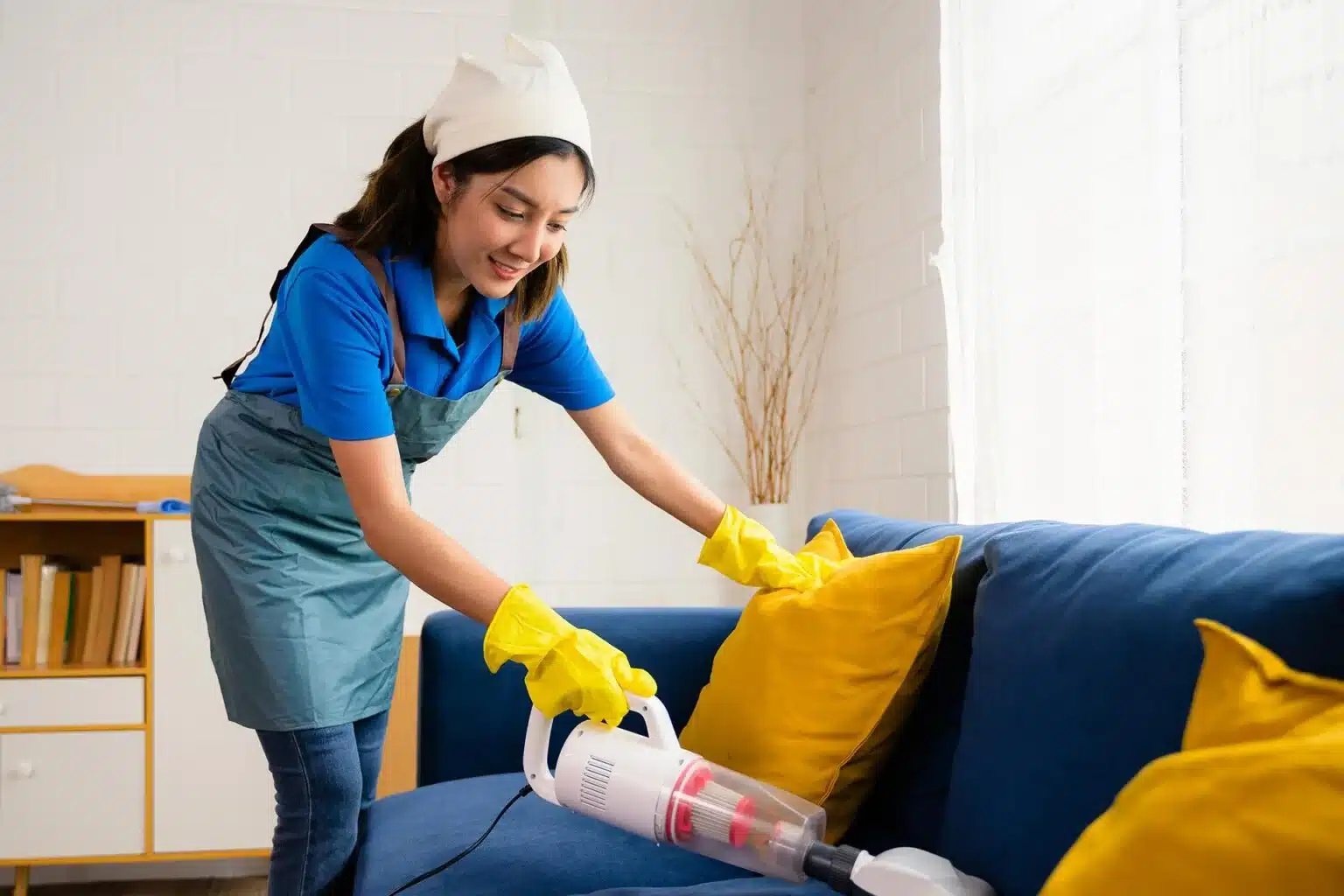 A smiling woman wearing gloves and an apron uses a vacuum cleaner to clean a blue sofa with yellow pillows in a bright living room.